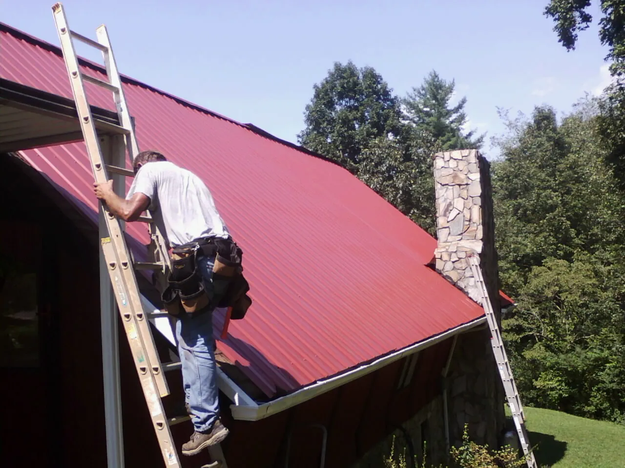 Roofer on a ladder beside a red metal roof and stone chimney — Soffit & Fascia Repair by Roofing Brothers in Forney